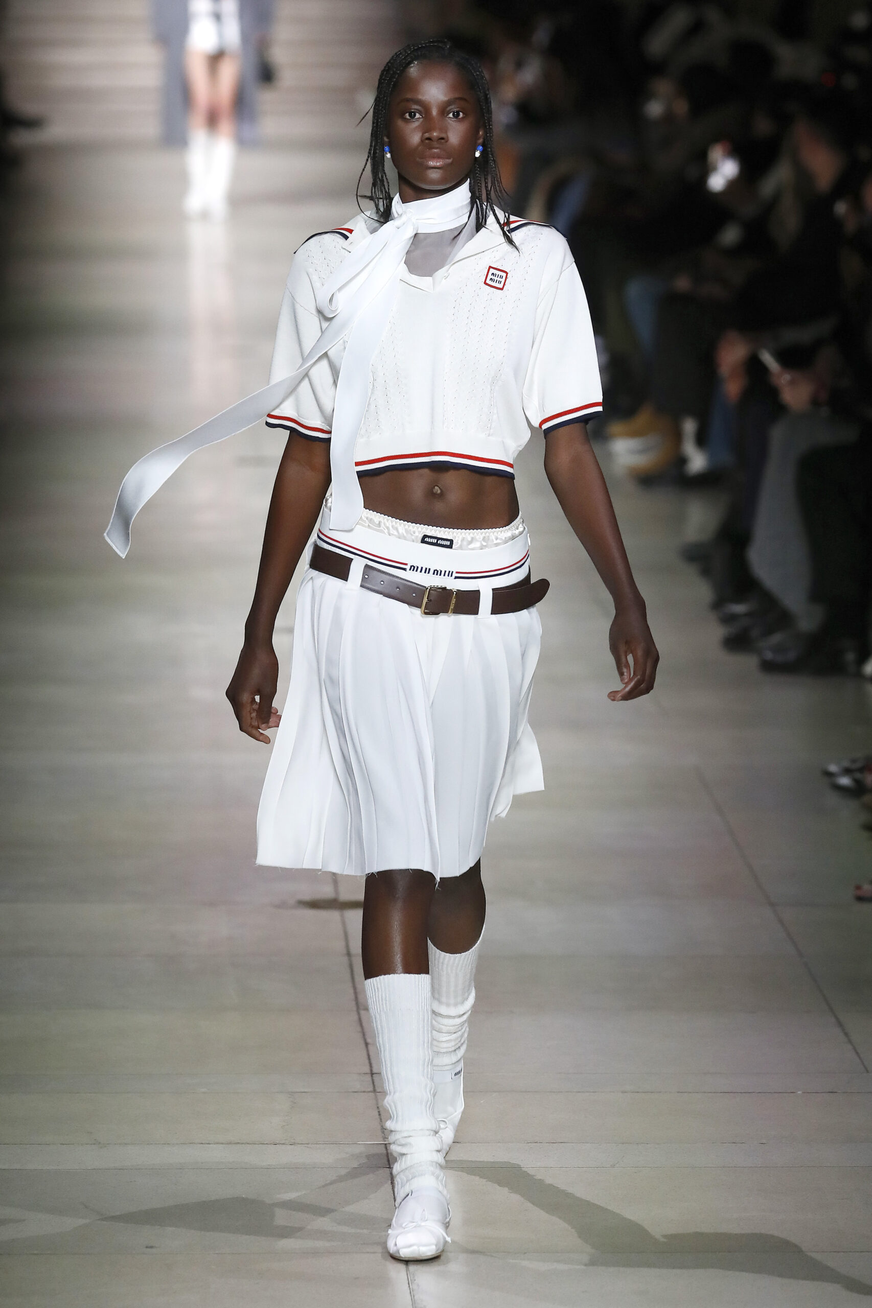 Egy modell fehér felsőben, szoknyában és fehér balerinában sétál a Párizsi Fashion Week kifutóján