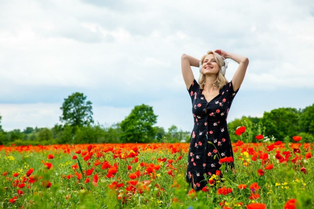 Nő hosszú fekete ruhában, virágokkal. A fűben áll a piros pipacsok között. Fejhallgató van a fülében