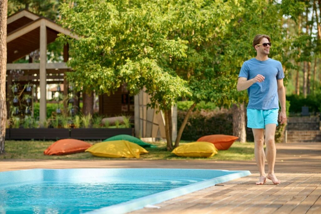 Egy fiatalember sétál a medence mellett, türkizkék fürdőnadrágot és kék pólót visel