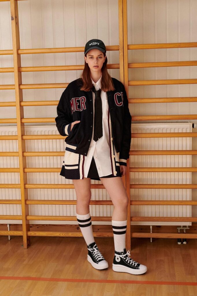 Egy lány baseballsapkában, egyetemi stílusú dzsekiben, rakott szoknyában, magas zokniban és sneakerben áll a bordásfal előtt
