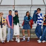 Egy csapat gyerek színes, különböző stílusú ruhában áll egy kapuban az iskola udvarán