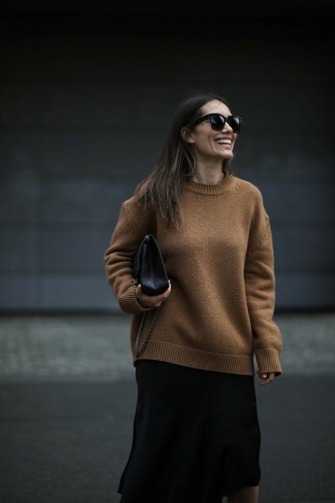 Egy nő barna pulóverben és fekete napszemüvegben, kezében egy fekete táskával