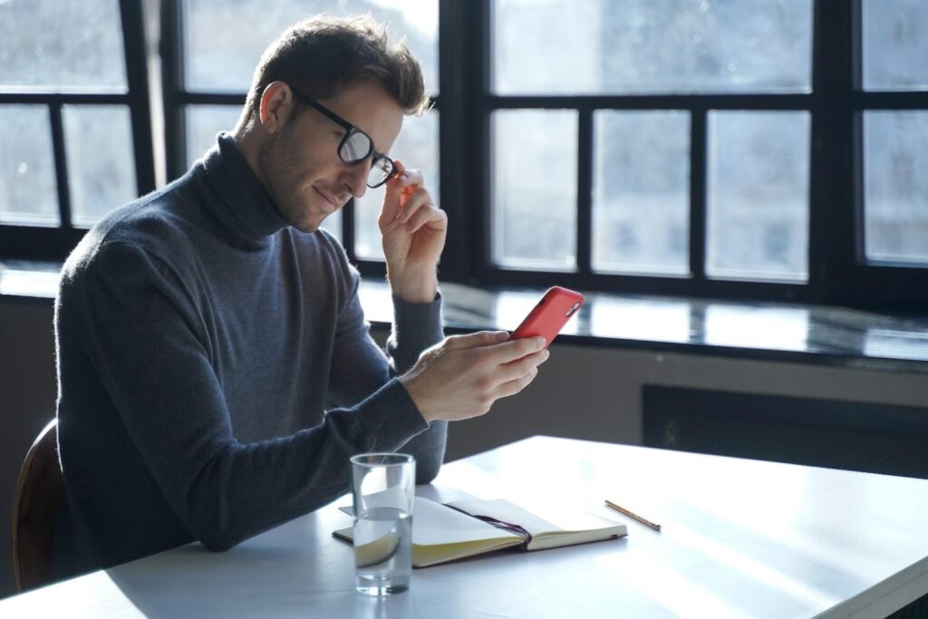 Egy fekete garbóba öltözött férfi ül egy íróasztalnál, kezében egy telefonnal